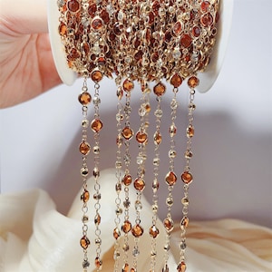 May include: A gold-toned metal chain spool featuring alternating round amber-colored and clear crystal-like stones set in bezel settings. The delicate chain hangs in multiple vertical strands from the white plastic spool, showcasing a repeating pattern of metallic links and faceted circular gems.