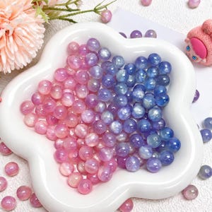 Peut inclure: Une collection de perles rondes et polies présentant un dégradé de couleurs allant du rose tendre au violet et au bleu profond. Les perles sont disposées dans un plat en céramique blanc en forme de fleur, avec quelques perles éparpillées autour.