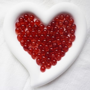 May include: A heart-shaped dish filled with red carnelian beads. The beads are smooth and polished, and they have a natural, earthy look.
