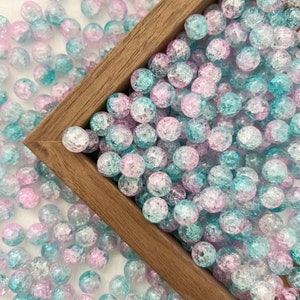 May include: A wooden box filled with a variety of small, round, iridescent beads. The beads are a mix of light blue and pink colors with a textured, frosted finish.