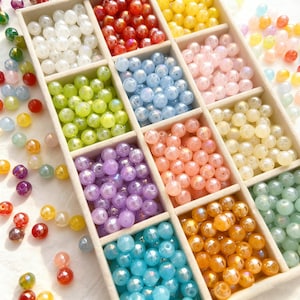 May include: A wooden organizer filled with colorful, iridescent beads. The beads are arranged by color in separate compartments, including white, red, yellow, green, blue, pink, orange, and purple. Some beads are scattered outside the organizer.