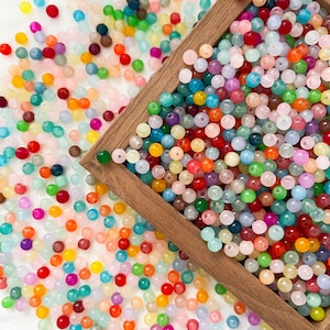 May include: A selection of multi-coloured, round beads in a variety of sizes and hues, including red, orange, yellow, green, blue, and purple. Some beads are in a wooden tray, while others are scattered. The beads appear to be made of glass or a similar material.