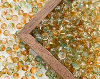 May include: A close-up of a wooden box filled with small, faceted glass beads. The beads are a mix of green and amber colors, creating a beautiful and unique look.