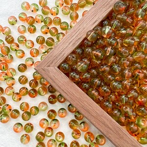 May include: A wooden frame filled with green and orange glass beads. The beads are scattered around the frame on a white surface.