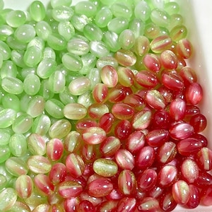 May include: A collection of oval-shaped glass beads featuring a smooth, polished finish. The beads are colored in shades of light green and deep red, with many displaying a subtle, shimmering gold glitter effect embedded within the translucent material.