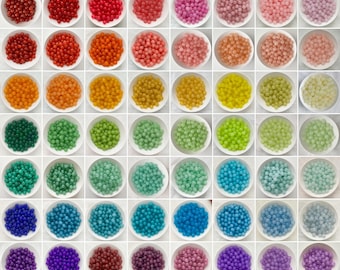 May include: A grid of 56 white bowls, each filled with a collection of small, round, smooth beads in a wide spectrum of colors. The beads are arranged in a gradient pattern, transitioning through shades of red, orange, yellow, green, blue, and purple.