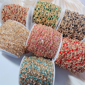 Könnte beinhalten: Sieben Spulen mit goldfarbenen Metallketten, die jeweils eine durchgehende Reihe von facettierten Steinen in den Farben Orange, Grün, Schwarz, Transparent, Pink, Rot und Hellblau aufweisen, angeordnet in einer Gruppe auf weißem Grund.