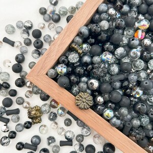 May include: A wooden box filled with a variety of black and grey glass beads. The beads are of different shapes and sizes, and some have iridescent finishes. The box is sitting on a white surface.