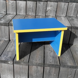 Puede incluir: Una pequeña mesa azul con un borde amarillo. La mesa está hecha de madera y tiene un diseño simple.