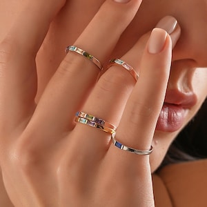 May include: A hand wearing five thin gold rings with colourful gemstones. The rings are stacked on two fingers.