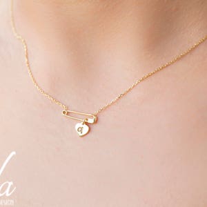 May include: A gold chain necklace with a safety pin charm and a small heart pendant with the letter 'a' engraved on it.
