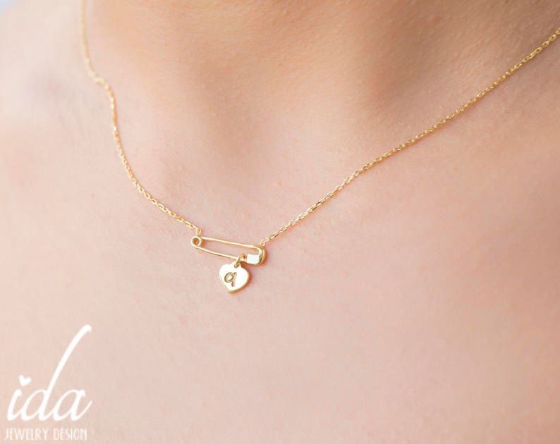 May include: A gold chain necklace with a safety pin charm and a small heart pendant with the letter 'a' engraved on it.