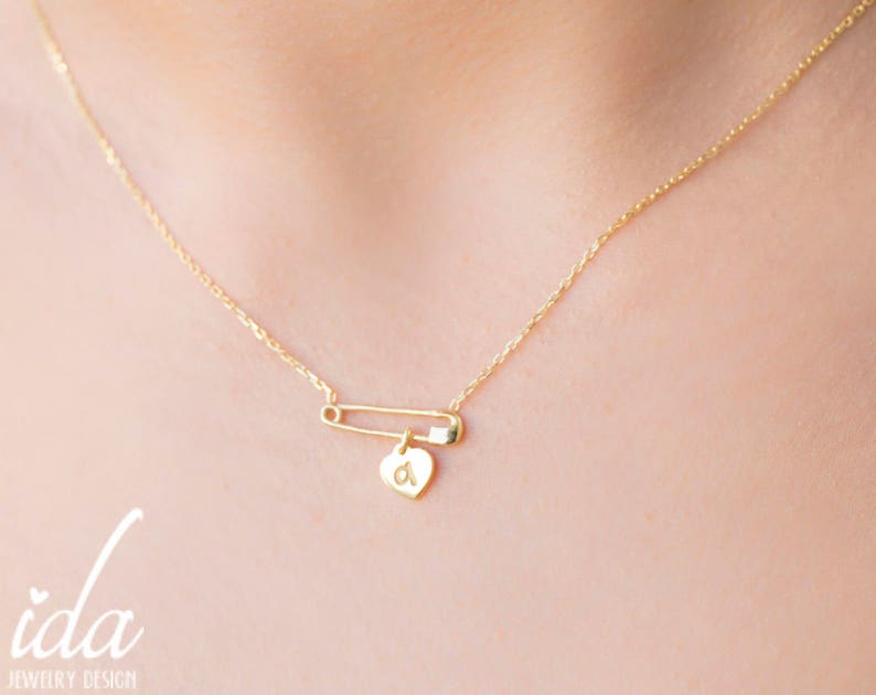 May include: A gold chain necklace with a safety pin charm and a small heart pendant with the letter 'a' engraved on it.