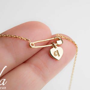 May include: A gold chain necklace with a safety pin charm and a heart pendant with the letter 'a' on it.