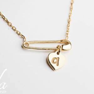 May include: A gold necklace with a safety pin charm and a heart pendant with the letter "q" engraved on it.