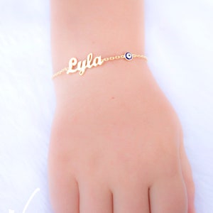 May include: Gold chain bracelet with the name "Lyla" and a blue evil eye charm.