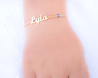 May include: Gold chain bracelet with the name "Lyla" and a blue evil eye charm.