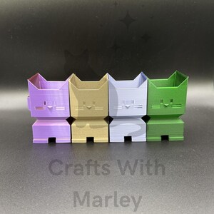 May include: Four small, blocky, cat-shaped containers in purple, gold, light blue, and green. Each figure features a simple geometric face with a triangular nose and horizontal line whiskers. The text Crafts With Marley is printed below.