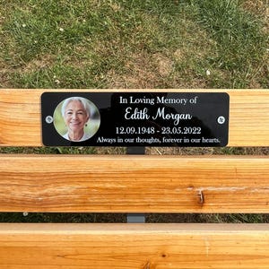 May include: A black memorial plaque fixed to a wooden bench. The plaque has a photo of a woman and the text "In Loving Memory of Edith Morgan 12.09.1948-23.05.2022 Always in our thoughts, forever in our hearts."