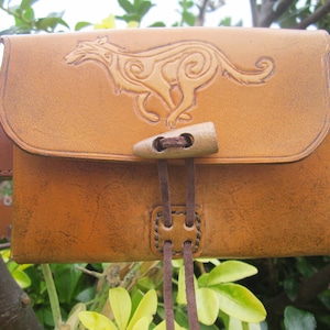 May include: A brown leather belt bag with a tooled design of a running wolf. The bag has a flap with a wooden button closure and two brown leather straps.