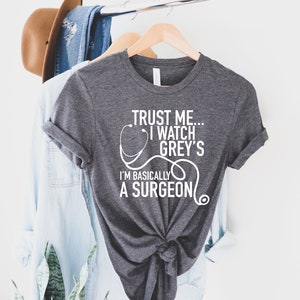 Puede incluir: Una camiseta gris oscuro con un gráfico blanco que dice "Trust me... I watch Grey's I'm basically a surgeon" con un gráfico de estetoscopio.