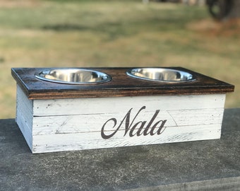 May include: A rectangular pet feeder stand with a dark brown stained wooden top and a white distressed wooden base. The front features the name Nala in black script. Two circular stainless steel bowls are set into the top surface.