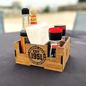 May include: A wooden condiment caddy with a wood grain finish, featuring a black circular logo that reads FILIPPS CAFE EST. 1951 A TRADITION CONTINUES. The caddy holds white paper napkins, a tall glass bottle, and two small spice shakers with red lids.