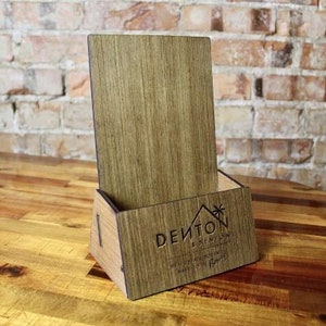 May include: A wooden brochure holder with a stand. The holder has a brown wood finish and the stand is made of a darker wood. The holder has the text "DEITON" and "EST. 2018" engraved on the front.
