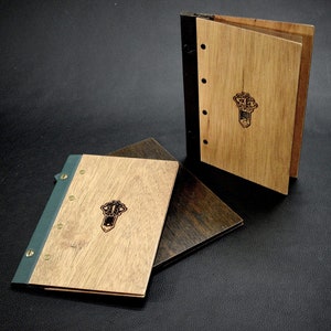 May include: Two wooden-cover notebooks with black spines and brass accents. The covers are a light wood with a dark wood inlay. Each notebook has a small, dark, engraved design on the front cover.