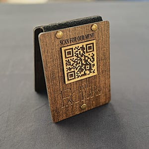 May include: A rectangular dark wood menu stand featuring a QR code and the text "SCAN FOR OUR MENU" and "THE FACTORY" engraved on the front. The wooden panel is secured with two brass-colored metal rivets at the top and bottom corners.