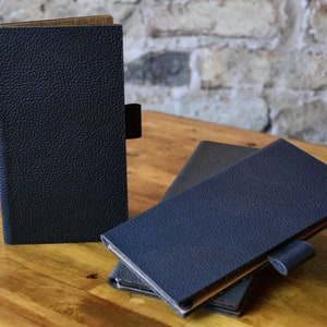 May include: Three blue leather-bound notebooks with a textured surface. The notebooks are stacked on a wooden table.