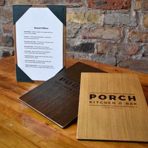 May include: Two rectangular wooden menu covers with the text PORCH KITCHEN & BAR engraved on the front, and one upright menu holder displaying a white paper dessert menu with black text, featuring items like cheesecake, tiramisu, and chocolate cake.