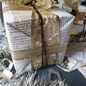 May include: A wrapped gift with a gold bow and ribbon. The wrapping paper is made from book pages, and the bow is adorned with gold filigree. Silver tinsel and a small tag that says "Merry Christmas" are in the foreground.