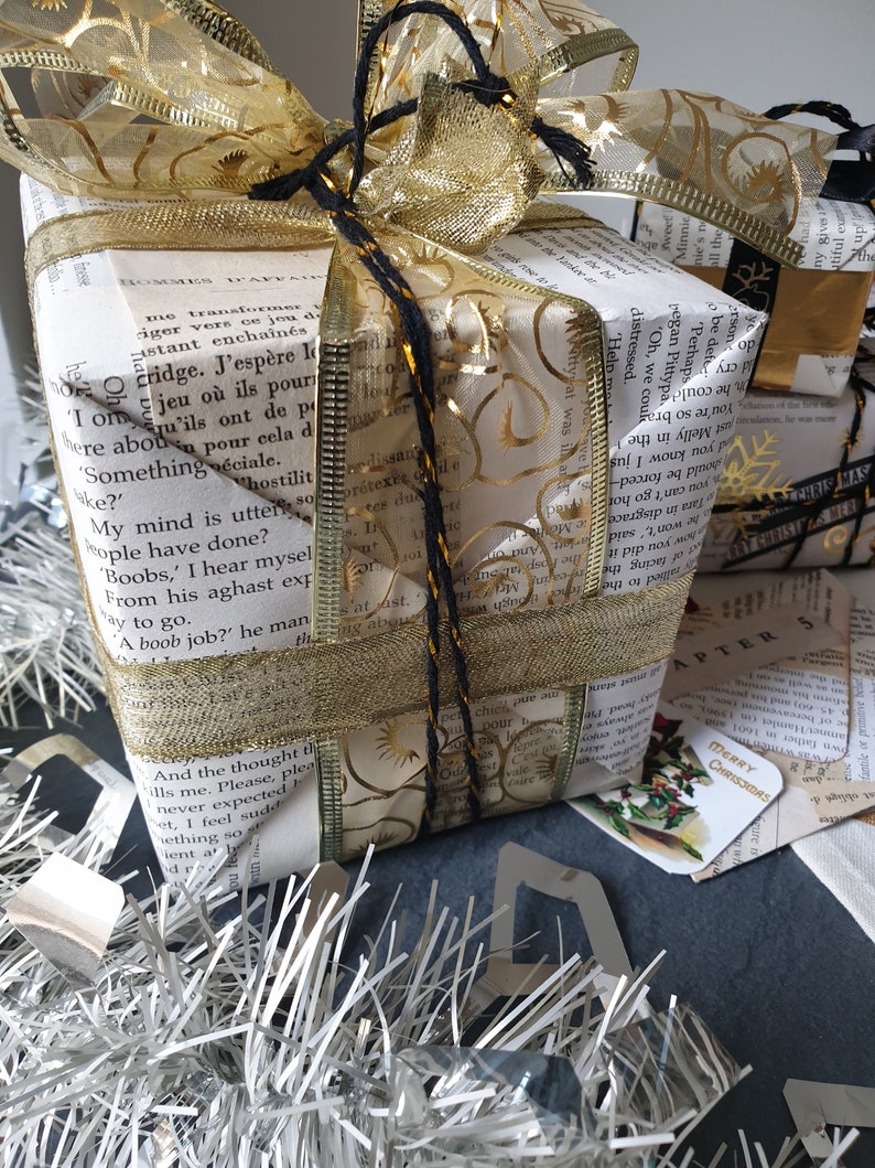 May include: A wrapped gift with a gold bow and ribbon. The wrapping paper is made from book pages, and the bow is adorned with gold filigree. Silver tinsel and a small tag that says "Merry Christmas" are in the foreground.