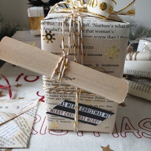 May include: A stack of wrapped gifts with a vintage book page design. The top gift is adorned with a white and gold ribbon, gold snowflake accents, and twine. A scroll and a "Merry Christmas" tag are also included.