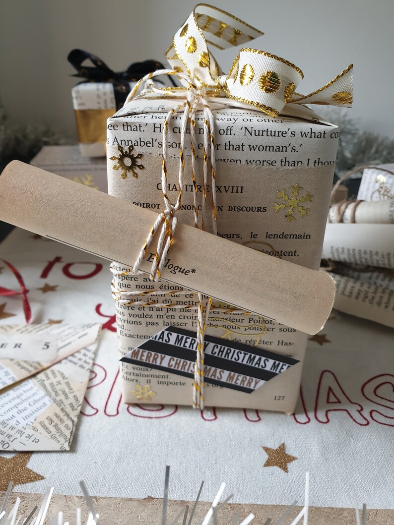 May include: A stack of wrapped gifts with a vintage book page design. The top gift is adorned with a white and gold ribbon, gold snowflake accents, and twine. A scroll and a "Merry Christmas" tag are also included.