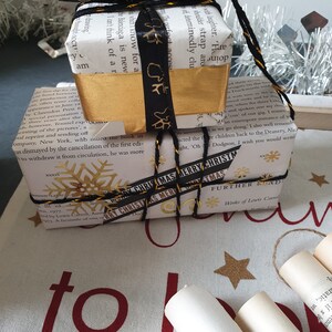 May include: Two wrapped gifts, one atop the other, are decorated for Christmas. The top gift is wrapped in book pages with a gold band and black ribbon. The bottom gift is wrapped in book pages with a gold snowflake and a ribbon that says "MERRY CHRISTMAS".