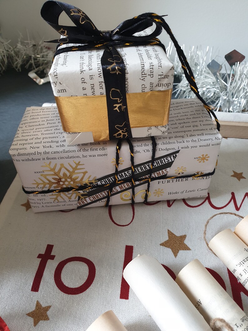 May include: Two wrapped gifts, one atop the other, are decorated for Christmas. The top gift is wrapped in book pages with a gold band and black ribbon. The bottom gift is wrapped in book pages with a gold snowflake and a ribbon that says "MERRY CHRISTMAS".