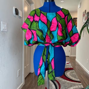 May include: A colorful top with a vibrant pattern of pink, teal, and green abstract shapes on a black background. The top is displayed on a blue dress form and has a matching tie belt.