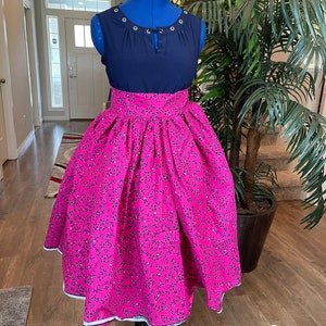 May include: A pink skirt with a floral print and a navy blue sleeveless top with a keyhole neckline. The skirt has a wide waistband and a gathered design. The top has a row of eyelets around the neckline.