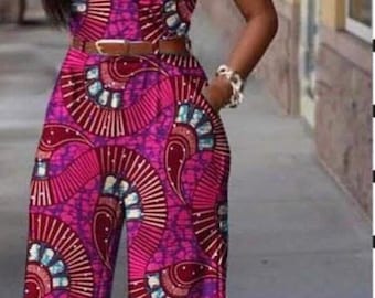 May include: A woman wearing a pink and purple jumpsuit with a geometric pattern. The jumpsuit has a V-neckline and a belt. The woman is standing in front of a brick wall with the word "BEAUTY" written on it.