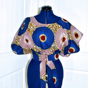 May include: A patterned top in blue, white, and red, with a matching tie belt, shown on a blue dress form. The top has a round neckline and short sleeves. The fabric features a circular design.