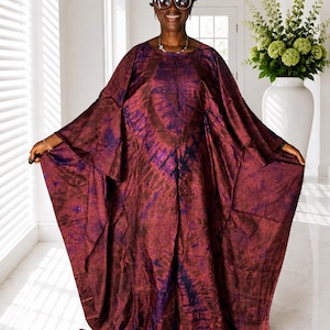 May include: A floor-length kaftan dress featuring a deep burgundy and dark purple tie-dye pattern. The garment has a loose, flowing silhouette with wide, batwing-style sleeves, a round neckline, and a smooth, slightly lustrous fabric finish.