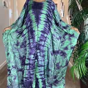 May include: A tie-dye caftan in shades of green and purple. The long-sleeved dress has a loose, flowing silhouette. The fabric appears soft and lightweight, suitable for warm weather. The caftan is a one-size-fits-all design.
