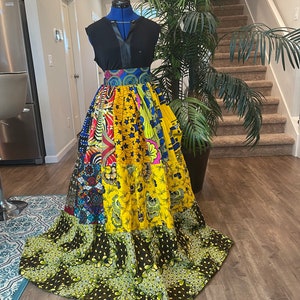 May include: A long, multi-colored patchwork skirt with a black top and a brown leather belt. The skirt is made of different fabrics with various patterns, including floral, geometric, and abstract designs. The skirt is gathered at the waist and flows to the floor.