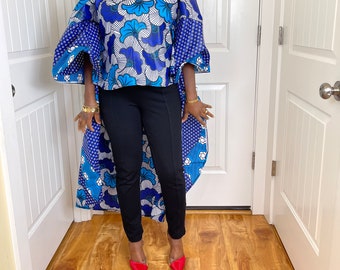 May include: A blue and white floral print off-the-shoulder top with a high-low hemline. The top has long sleeves with a gathered detail at the shoulders. The model is wearing black pants and red heels.