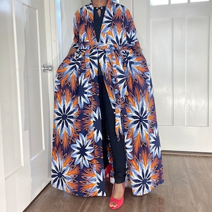 Op de afbeelding: Een lange, blauwe, witte en oranje bloemenprint-jas met een ceintuur en zijzakken. De jas is open aan de voorkant en heeft een lange, vloeiende rok.