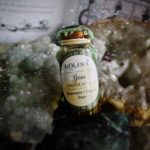 Könnte beinhalten: Eine kleine grüne Glasflasche mit einem Korkverschluss und einem weißen Etikett mit der Aufschrift "HOLDKØ Gone - Magick Bottle - Rosemary, Thyme, Sage". Die Flasche steht auf einem weißen und braunen Stein mit einem funkelnden Hintergrund.