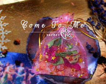 Op de afbeelding: Een roze gaas zakje met een zilveren pentagram bedel en de woorden "Come To Me sachet" erop gedrukt. Het zakje is gevuld met roze en groene kruiden en kristallen.