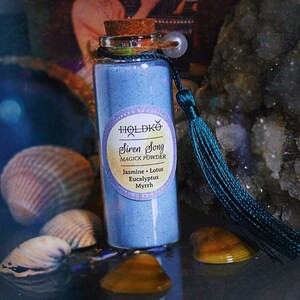 May include: A small glass bottle containing blue powder. The bottle has a cork stopper and a teal tassel. The label reads "Holdkö Siren Song Magick Powder Jasmine - Lotus Eucalyptus Myrrh".